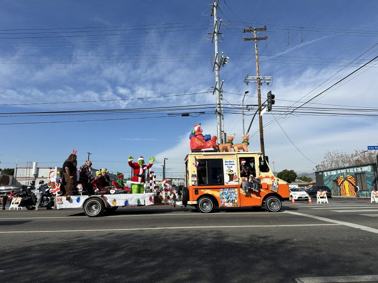 A wide shot of Boo Boo’s ice cream truck with a small float behind it. The ice cream truck is pale yellow and orange. On the roof is an inflatable Santa with a sleigh and reindeers. Behind, the float has children dressed as elves, and the Grinch waving at the crowd.