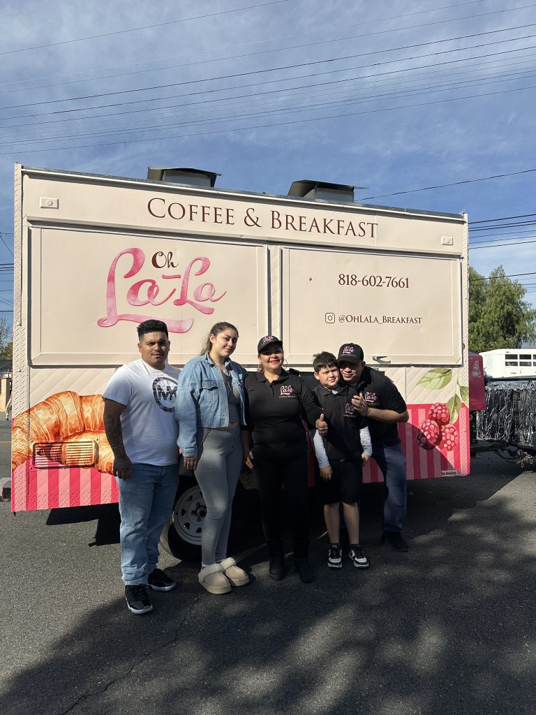 The family of the small business Oh La La Coffee and Breakfast group up to take a picture at the back of their food truck. The mother stands at the center with everyone around her. The back of the food truck has the name of the business and how to contact them. The bottom half of the food truck has vertical pink stripes across it. Above them is a blue sky with clouds.