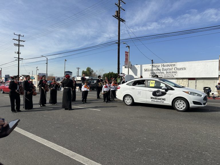 A wide shot of a small mariachi band. In front of them is their white car. To the left, the mariachi band prepares their instruments. To the right, children clothed in white dress shirts and black pants get ready. Behind them are more of the young adult members of the mariachi band.