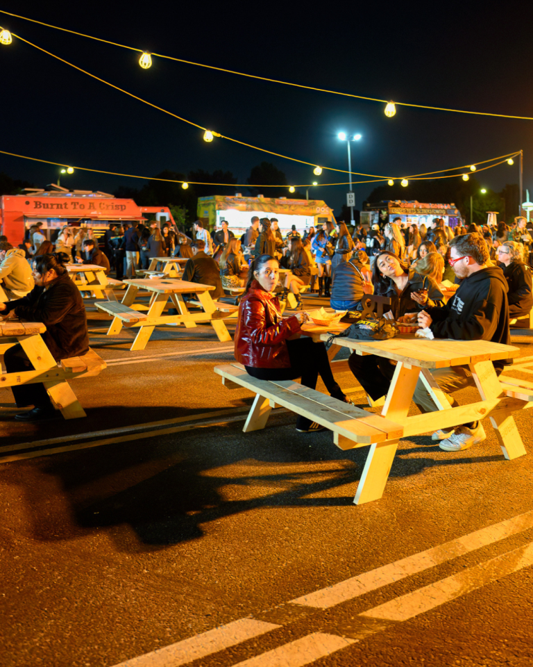 A crowd gathers at the food court to eat. In the distance, three food trucks cater to more guests.