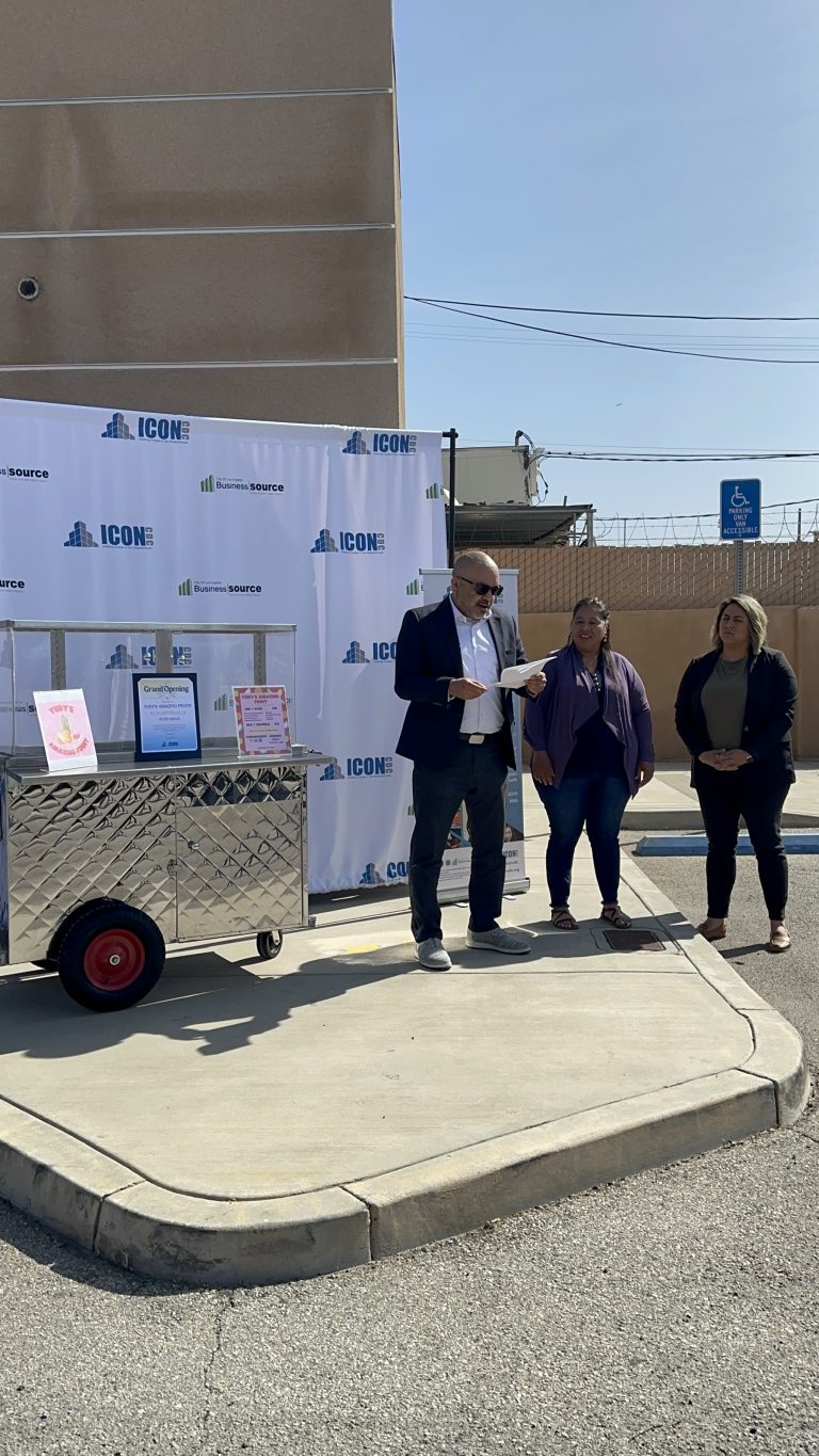 A wide shot of an ICON CDC representative giving a speech with the script in hand. Beside him are Yudy Mena and Senator Caroline Menjivar. To the other side is Rudy's fruit cart. Behind him is ICON CDC's backdrop and roll-up banner.