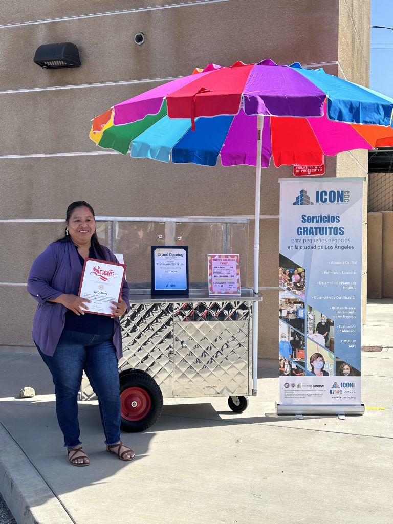 A wide shot of Yudy Mena holding her award. She stands beside her fruit cart that has her second award and the menu of her business. To the right is a roll-up banner of ICON CDC's information. In the back is a rainbow umbrella.