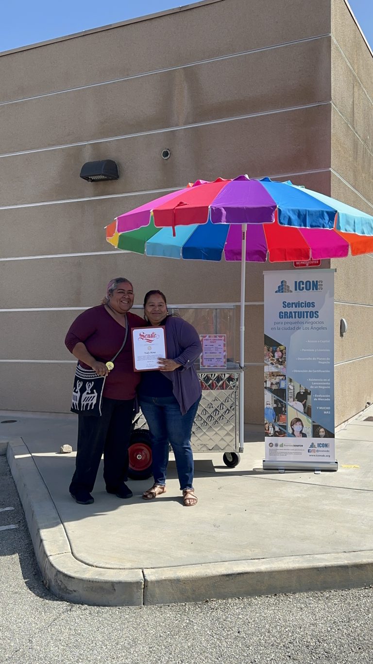 Wide shot of Yudy Mena posing for a photo with her award. Her relative is beside her. The fruit cart, rainbow umbrella, and ICON CDC roll-up banner is behind them.