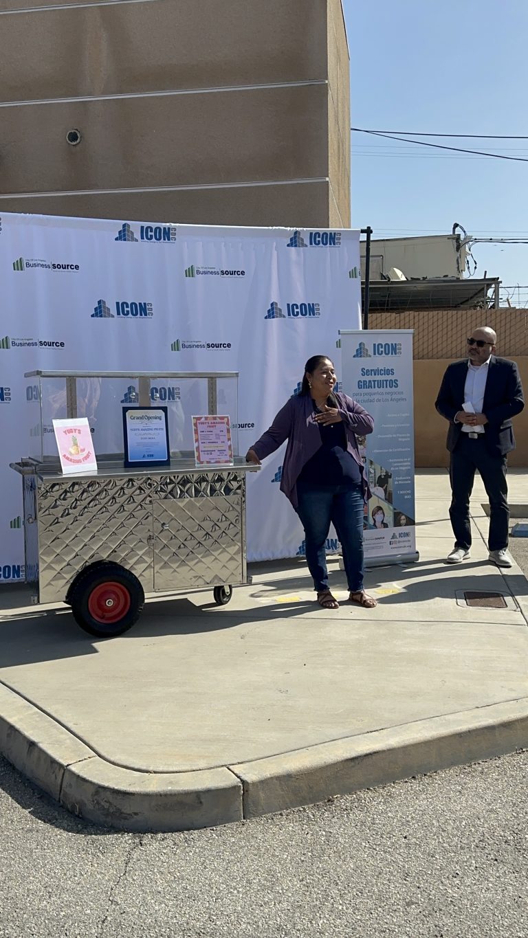 A wide shot of Yudy Mena with her hand against her chest as she gives a speech. Beside Yudy is her fruit cart. A few feet to the side is an ICON representative. Behind Yudy is the ICON CDC backdrop and roll-up banner.