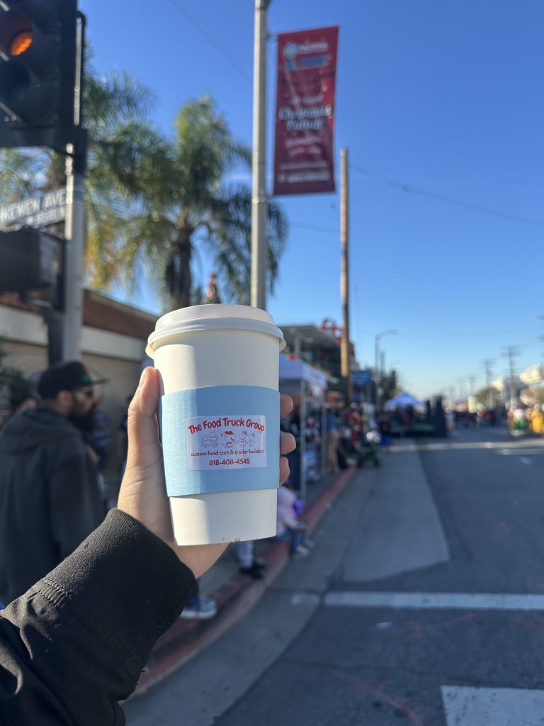 A hand holds a coffee cup with a sleeve that has The Food Truck Company’s name and phone number. Behind the hand is the street, and at the left is a sidewalk. The crowd huddles there, with some pop up canopy tents. Clear sky.