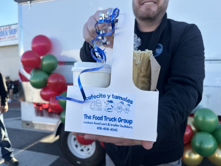 A wide shot of a smiling man holding a drink carrier. In the left carrier pocket is a coffee cup, in the right is a tamale. The carrier has a sticker with The Food Truck Group's name and phone number. Atop the carrier are silver and blue curling ribbons. Behind the man is a white food truck with balloons.