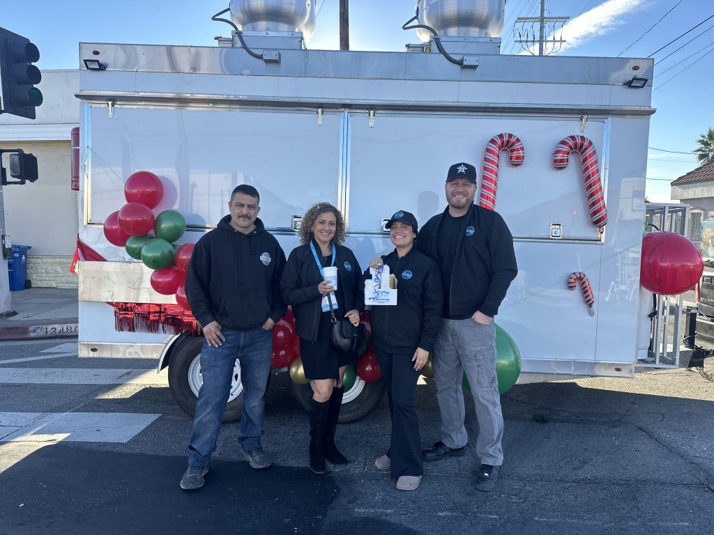 In a wide shot, four members of The Food Truck Company stand together for a picture. Maria Ponce and Kimberly Tapia are at the center, holding a coffee cup and the drink carrier containing a coffee cup and tamale. Behind the group is the white food truck with balloons. Above them is a clear blue sky.