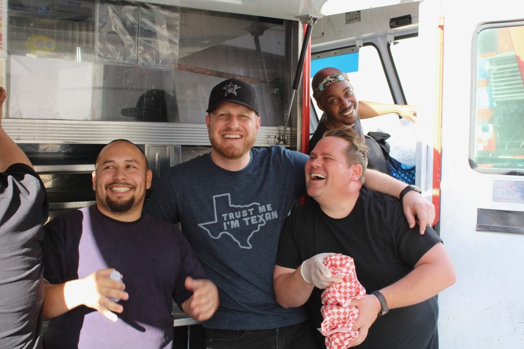 A wide shot of Burnt To A Crisp’s team close together for the camera. Everyone is smiling. One of the food truck’s owners has an arm around the second owner’s shoulder. Everyone stands beside the open service side window, close to the cabin.