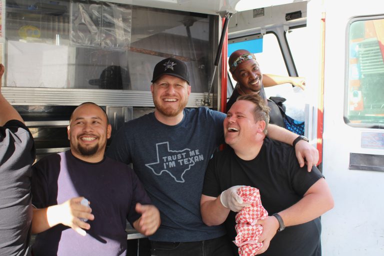 A wide shot of Burnt To A Crisp’s team close together for the camera. Everyone is smiling. One of the food truck’s owners has an arm around the second owner’s shoulder. Everyone stands beside the open service side window, close to the cabin.