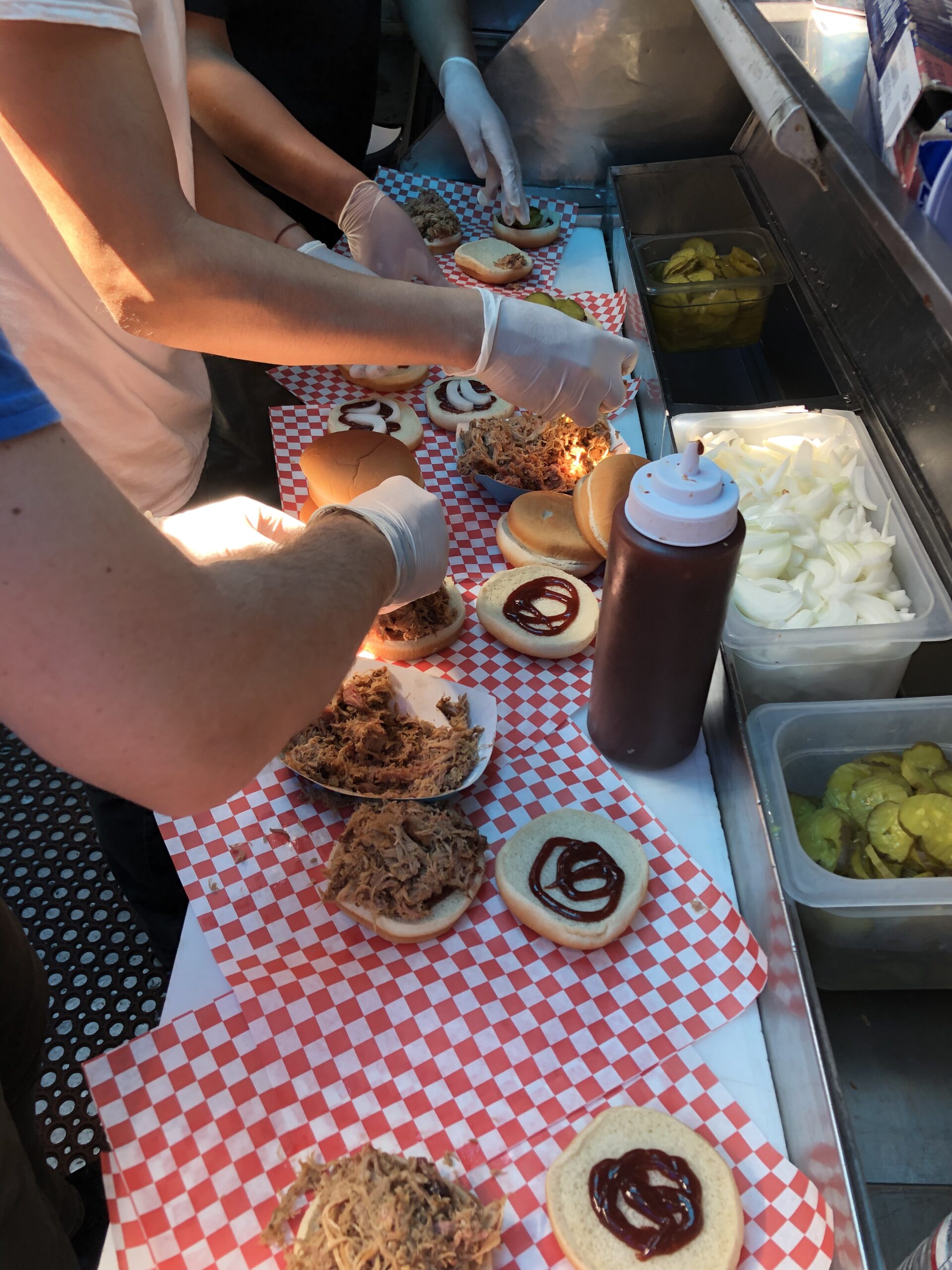 A vertical shot of chopped brisket hamburgers. Three workers stand close, hands covered in gloves. They spread chopped brisket from paper trays onto the hamburger buns. The condiment is spread over the top bun. Behind the condiment are containers of pickle slices and onions. Beneath the hamburgers is white and red checkerboard paper to wrap the complete burgers in.