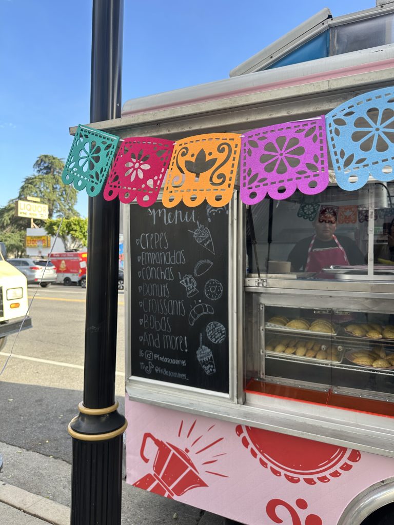 Wide shot of the left side of Frida’s Cravings food truck. The service side window is open, revealing pastries behind a glass window. Above the display is a baker inside the food truck. To the left of the service side window is a black chalkboard. White chalk outlines some of the menu, drawings of food, and the business’s instagram and TikTok handles. The background is of a street with a clear, blue sky.