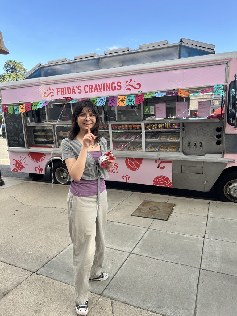 Wide shot of the service side of Frida’s Cravings. The service side window is open, showing pastries behind the glass. Papel picado hangs above. In front of the food truck is a female customer. Her right hand is raised, index finger up. In her left hand she holds tape.