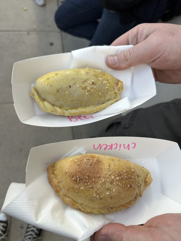Two paper food trays hold a single empanada each, vertically across from each other. The filling of the empanada is written on the side with a pink marker. The first is beef, and the second has chicken filling.