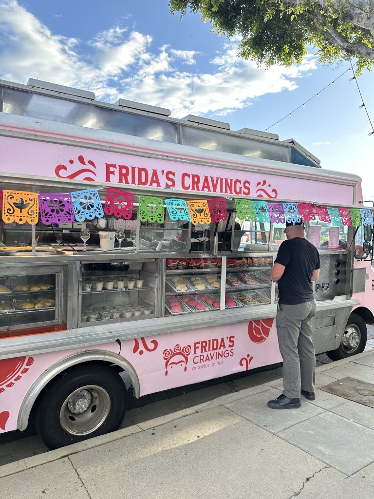 A vertical shot of Frida’s Cravings food truck with the service side window open. Pastries are visible behind the glass. Standing before the food truck is a male customer, looking at the pastries. The sky is blue with clouds overhead.