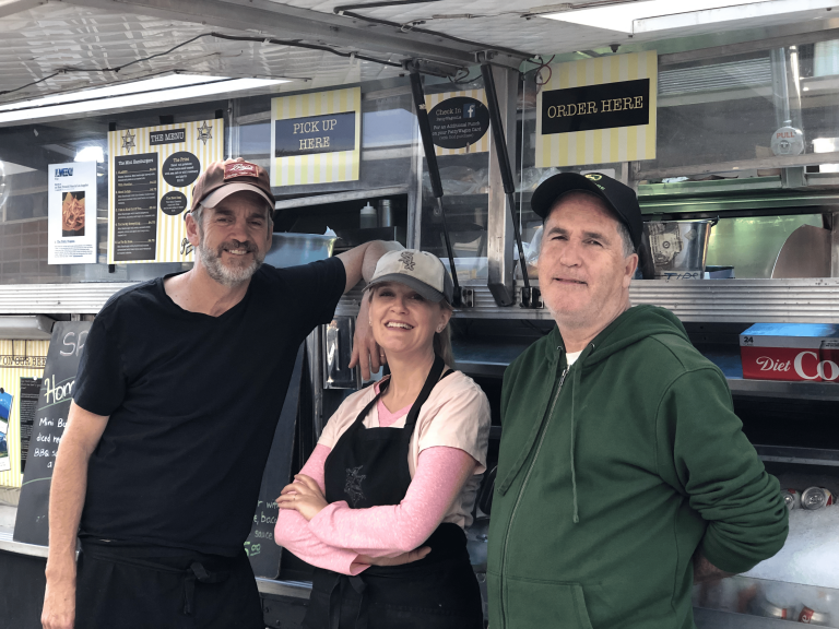 A wide shot of The Patty wagon from the open side view window. Directly in front of the food truck are the three owners, all standing together. The shot is closer, showing only the upper halves of the owners as they stand close together for the picture.