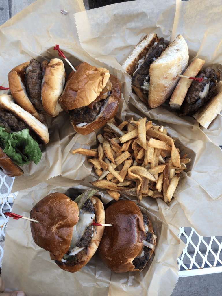 A portrait shot of The Patty Wagon’s hamburgers and fries. The seven hamburgers are atop paper trays with wax paper beneath them. In the center of all the hamburgers are a pile of fries, also inside a paper tray. They are all set on a white table outside.